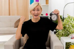 excited-beautiful-blonde-russian-woman-sits-armchair-keeping-fist-holding-alarm-clock-inside-living-room